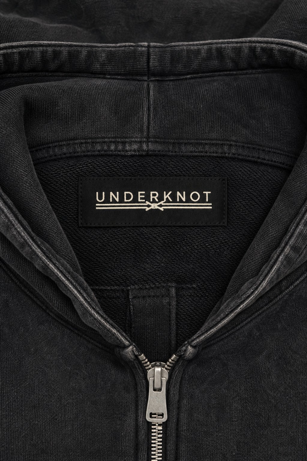 HEAVY HOOD ZIP-UP 넥 안쪽 중앙의 블랙 직조 UNDERKNOT 메인라벨과 웜 에크루 엠블럼을 보여 주는 디지털 디테일. 사이즈 표기 없음