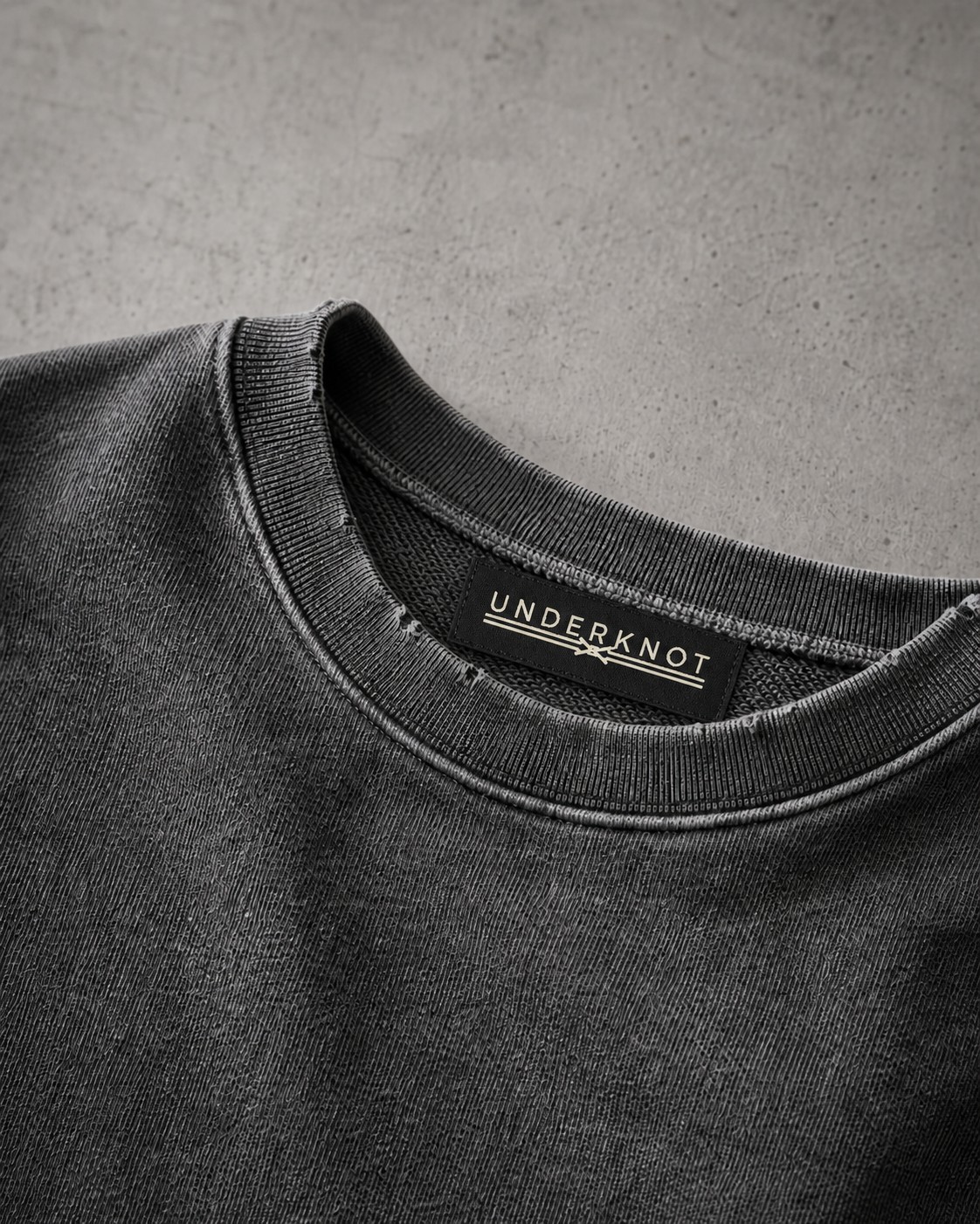 FADED GRAPHITE 넥 안쪽 중앙의 블랙 직조 UNDERKNOT 메인라벨과 웜 에크루 엠블럼을 보여 주는 디지털 디테일. 사이즈 표기 없음