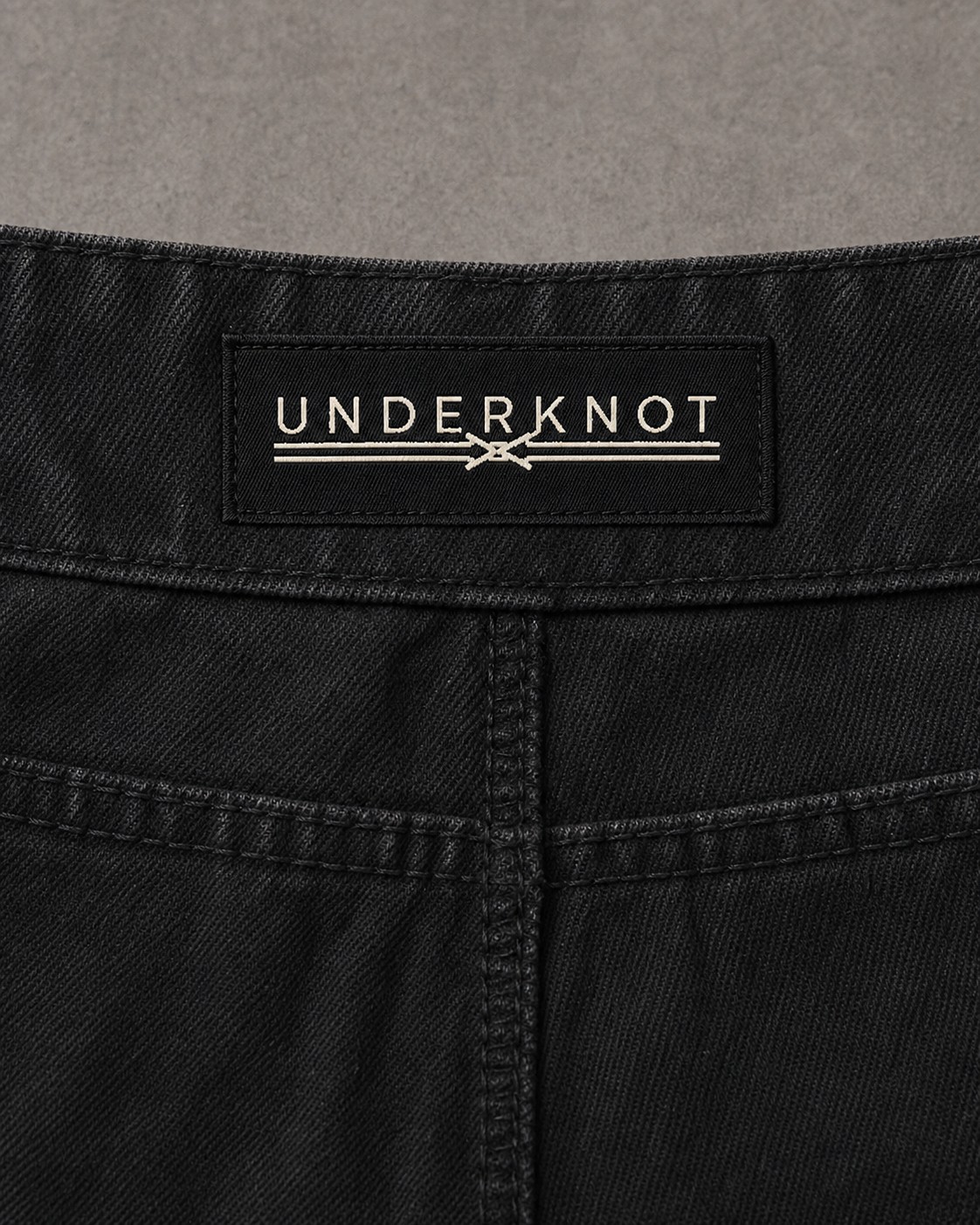 WASHED BLACK 허리 안쪽 중앙의 블랙 직조 UNDERKNOT 메인라벨과 웜 에크루 엠블럼을 보여 주는 디지털 디테일. 사이즈 표기 없음